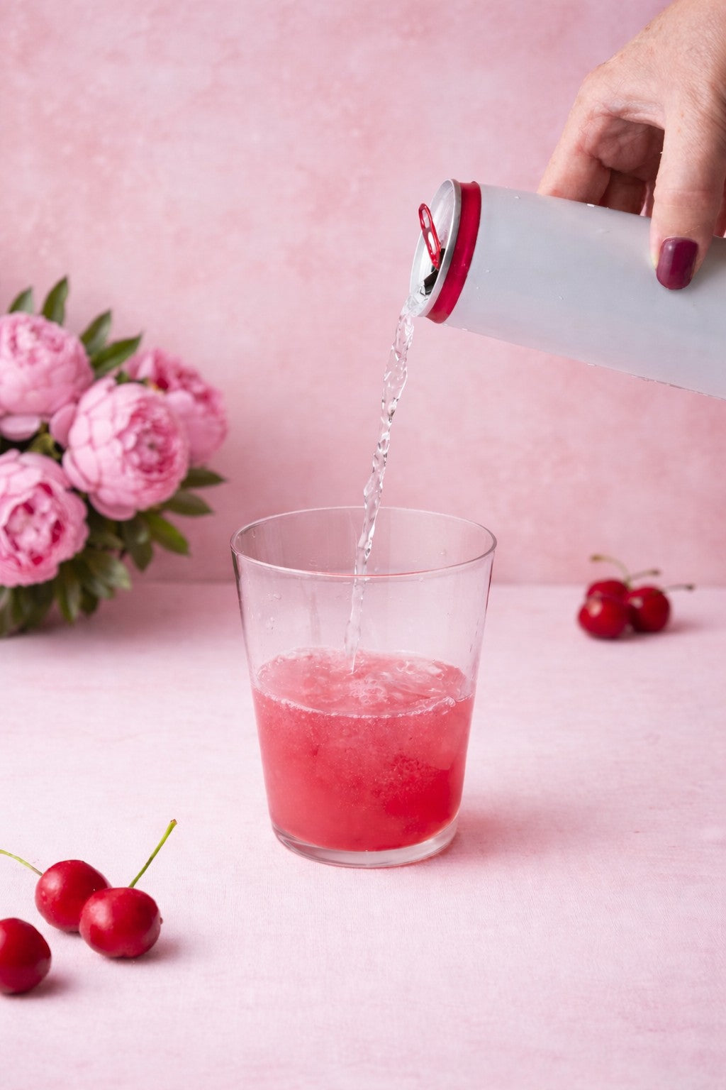 Pink drink being poured into a glass with cherries and flowers on a pink background