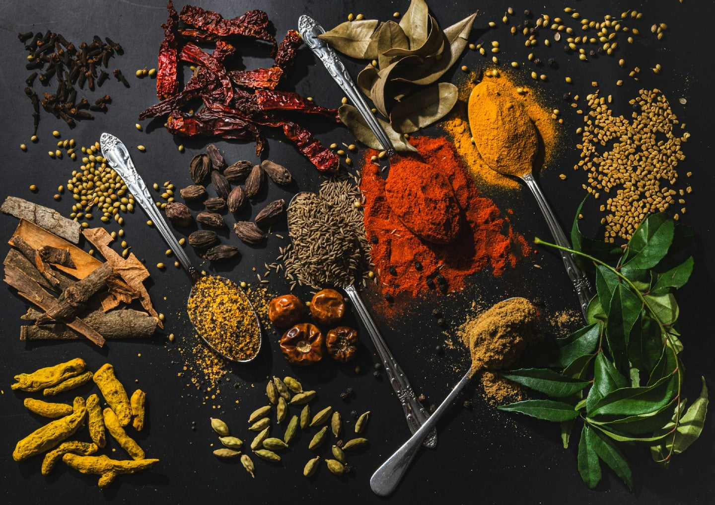 Assorted spices and herbs on a dark surface with silver spoons.